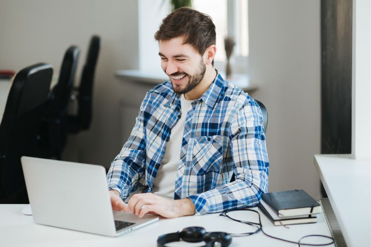 Entrepreneur engaged in a leadership training session, smiling while working on a laptop at a modern desk. The workspace includes notebooks and headphones, suggesting focus and readiness for strategic growth. The setting conveys a casual yet professional environment ideal for small business development.
