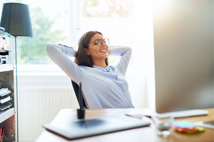 Team member enjoying a moment of relaxation during a workplace wellbeing session, seated at a desk with hands behind the head and smiling. The bright, modern office setting—with computer, notes, and natural light—reflects a supportive environment that encourages balance, mental clarity, and positive team culture.